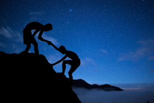 Silhouettes of climbers helping one another up a mountain at night.
